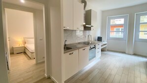 Una cocina moderna con armarios blancos, un lavamanos de acero inoxidable y un horno empotrado. Se ve una ventana con vistas a edificios.