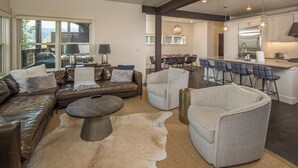 A spacious living area with a leather sofa, a round coffee table, and a light-colored armchair. A dining area with a bar stool and a kitchen with stainless steel appliances is visible in the background. - New! Hot Tub, Soaking Tub, Great Location! (Big Sky)