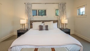 A large bed with a wooden headboard, two bedside tables with lamps, a bench at the foot of the bed, and two windows with curtains. - New! Hot Tub, Soaking Tub, Great Location! (Big Sky)