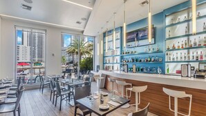 A modern restaurant with a bar area, large windows with a view of palm trees and buildings, and a variety of bottles on shelves. - Studio with Garden View and Balcony – 3rd Floor (Hallandale Beach)