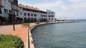 A waterfront promenade with a paved walkway, a building with arched windows, and a calm body of water. - Air-conditioned apartment with splendid panoramic view - COUDALERE pedestrian village (barcares)