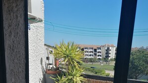 A balcony with white cushioned seating, a potted plant, and a view of buildings in the background. - Air-conditioned apartment with splendid panoramic view - COUDALERE pedestrian village (barcares)
