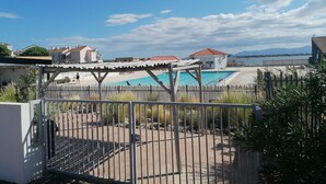 A pool area with a wooden deck, a fenced pool, and buildings in the background. - Air-conditioned apartment with splendid panoramic view - COUDALERE pedestrian village (barcares)