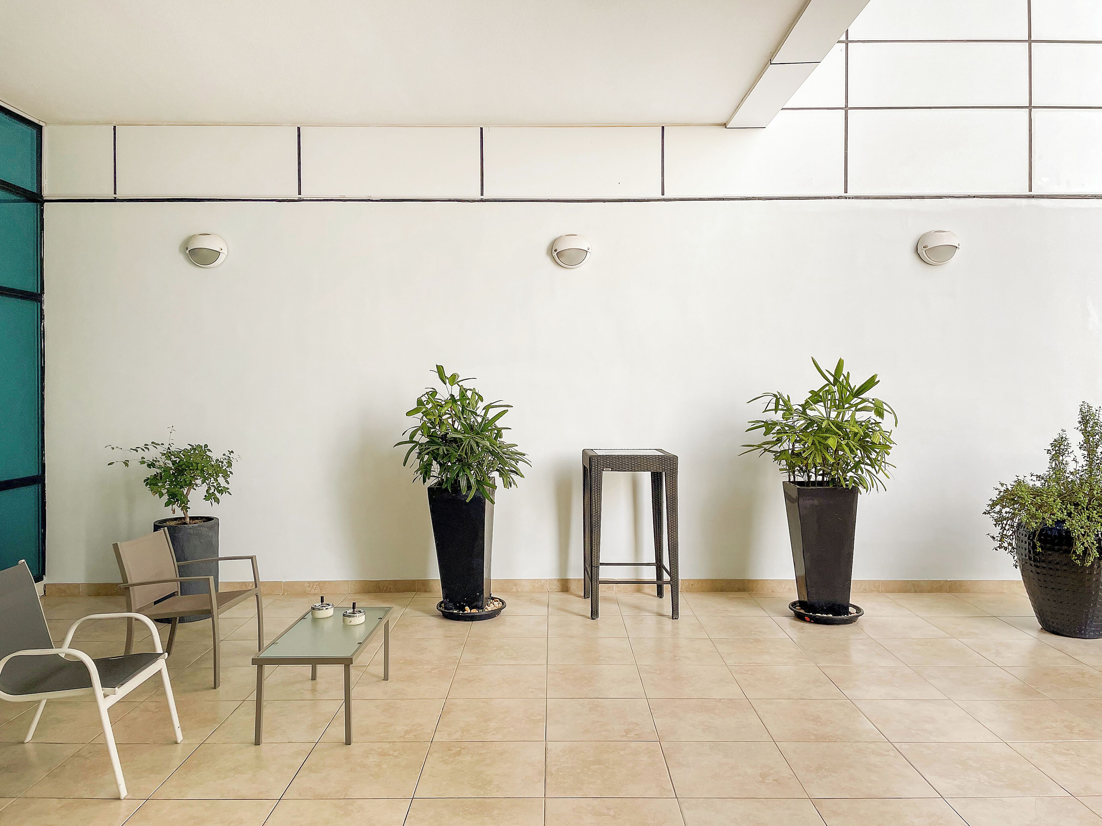 A modern waiting area with a tiled floor, a glass table, two chairs, and four potted plants.