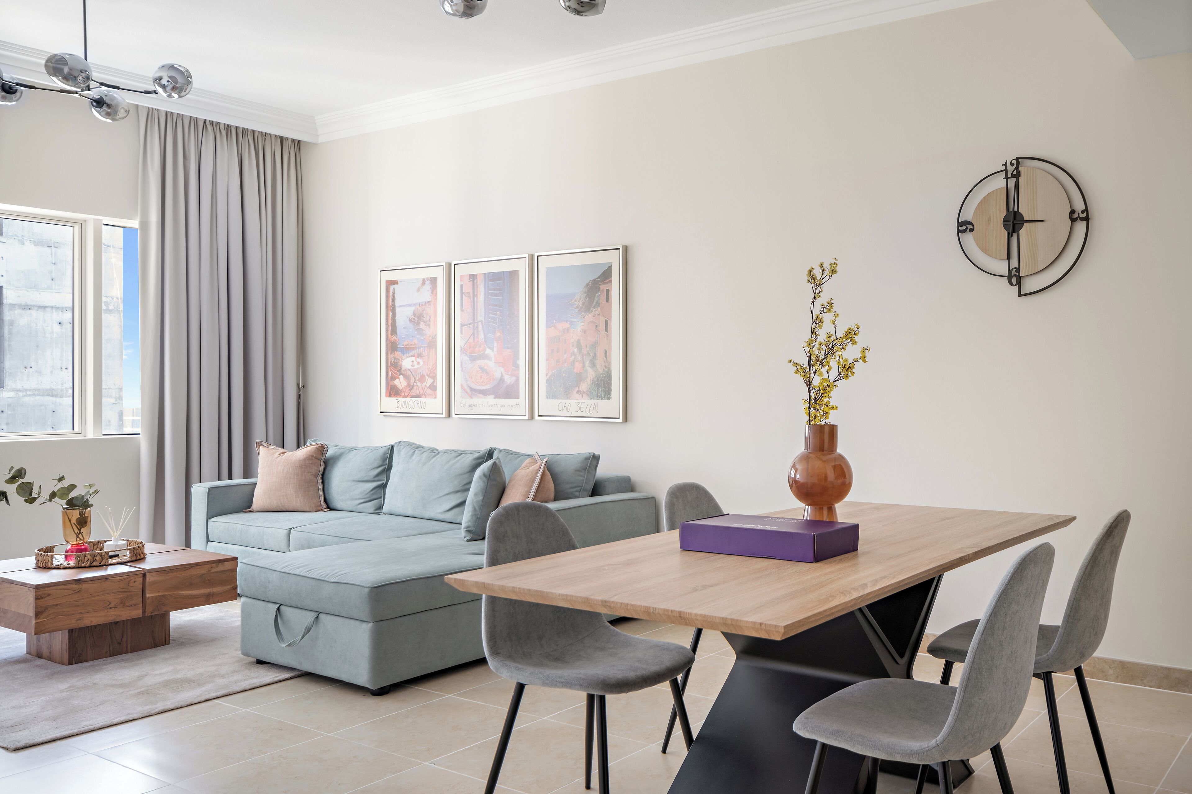 A modern living room with a sofa, a dining table with chairs, a vase with flowers, and a wall clock.