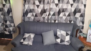 Un sofá gris con dos cojines estampados, una cortina con diseño y una mesita con un cubo de Rubik y un objeto blanco.