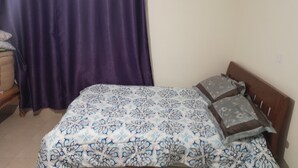 Un dormitorio con cama, cabecera de madera, cortina morada y una cesta tejida en el suelo.