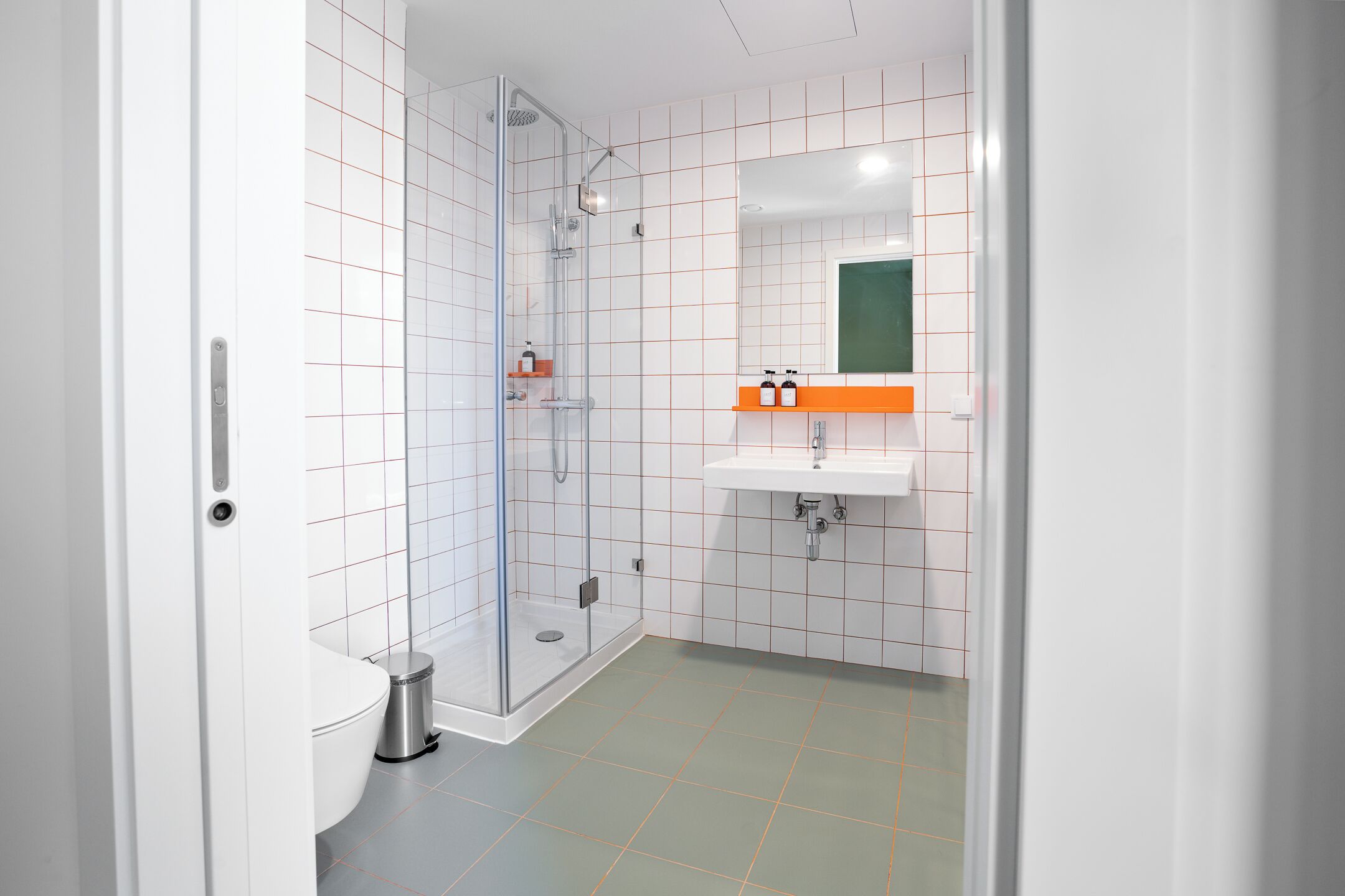 Un baño moderno con cabina de ducha de vidrio, un lavamanos blanco y un estante naranja, y un espejo sobre el lavamanos.