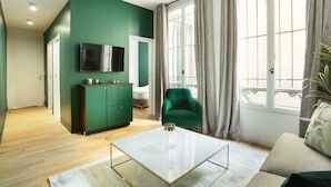 A modern living room with a green accent wall, a flat-screen TV mounted above a dark green cabinet, a green armchair, and a beige sofa with a decorative pillow.