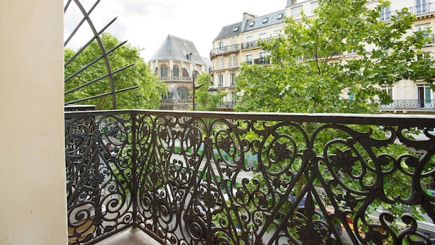 A balcony with ornate wrought iron railings overlooking a street with historic buildings and lush greenery.