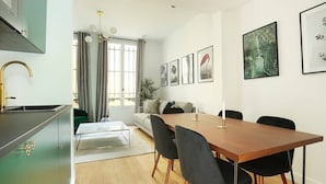 A modern kitchen with a stainless steel range hood, a dining area with a wooden table and black chairs, a sofa with a green cushion, and a large window with curtains.