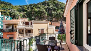 A balcony with a glass table and chairs, a striped umbrella, and a view of a hillside with buildings. - Apartment 'Ce Tour - Casa Cetara Centro' with Balcony, Wi-Fi and Air Conditioning (Cetara)