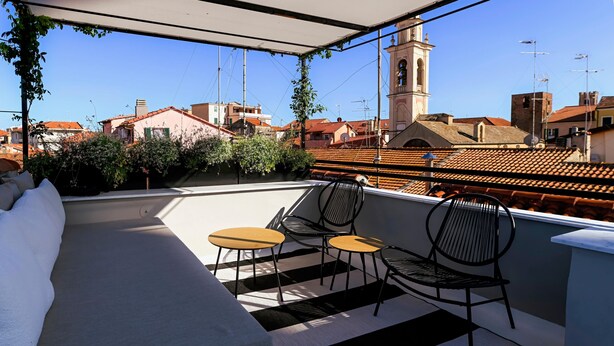 A rooftop terrace with a sofa and two small tables, surrounded by a railing with a view of buildings and a tower. - ‘Torri e Tramonti’ Apartment with Private Terrace and Air Conditioning (Albenga)