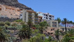 A coastal residential area with multi-story buildings, some with balconies, and lush greenery including palm trees. - Apartamentos Gran Rey Nº 8 (Valle Gran Rey)