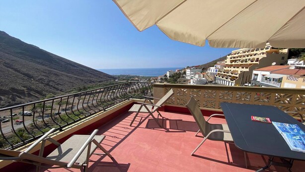 A balcony with a table and chairs, overlooking a coastal road and buildings. - Apartamentos Gran Rey Nº 8 (Valle Gran Rey)