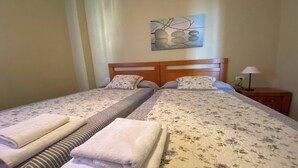 A double bed with floral bedding, two pillows, and a wooden headboard. There is a nightstand with a lamp and a wall-mounted artwork above the bed. - Apartamentos Gran Rey Nº 8 (Valle Gran Rey)