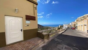 A narrow street with a white door and a sign indicating a room number. The street leads to a coastal view with buildings and a clear sky. - Apartamentos Gran Rey Nº 8 (Valle Gran Rey)