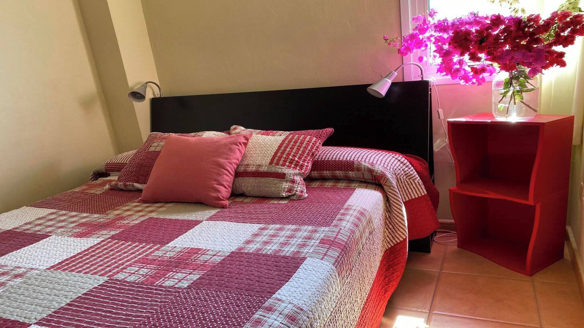 Una cama bien hecha con una colcha a cuadros, un cabecero negro y una mesita de noche roja con un jarrón de flores.