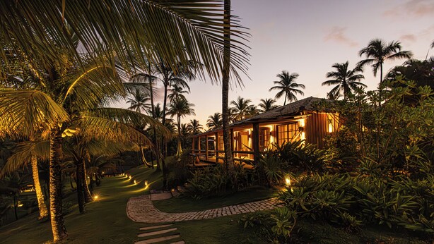 A tropical resort at dusk with a well-lit pathway leading to a wooden building surrounded by palm trees and lush greenery. - Fazenda Jeribucaçu (Itacaré)
