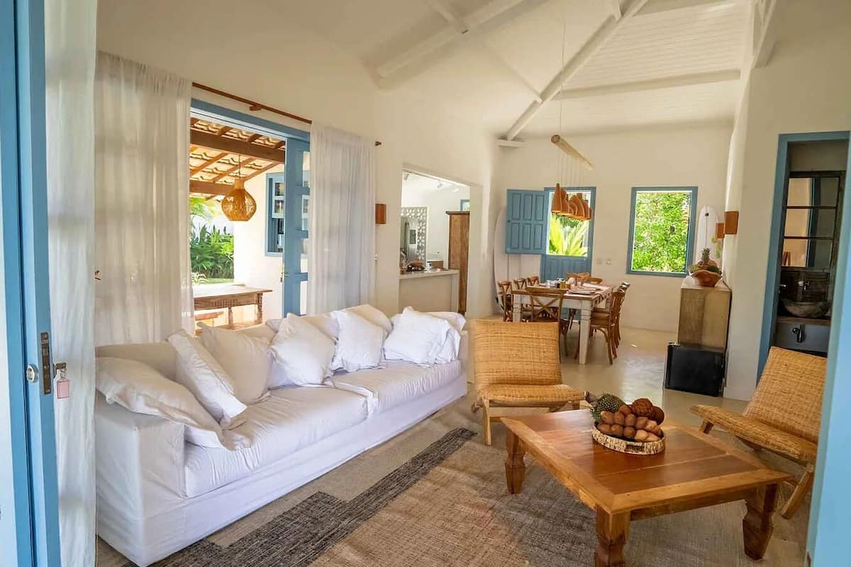 A spacious living area with a white sofa, a wooden coffee table with fruits, and a dining area with blue shutters.