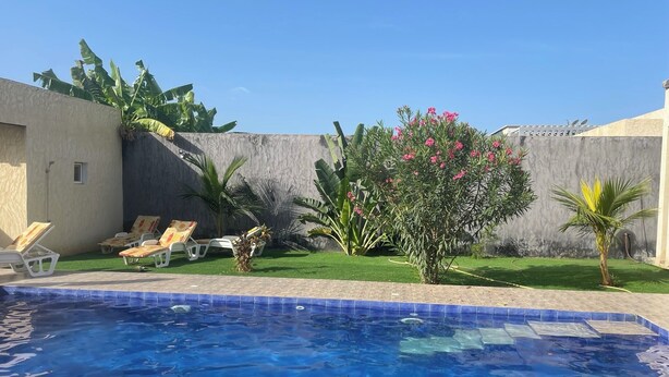 A swimming pool with a tiled edge, surrounded by a grassy area with palm trees and a small bush with pink flowers. There are lounge chairs along the poolside. - Luxury villa with private pool and staff close to the beach and Saly (N'Guerigne Bambara)