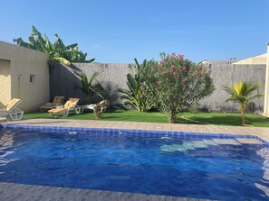 Une piscine dont le bord est revêtu de carreaux, entourée d’une pelouse agrémentée de palmiers et d’un petit arbuste aux fleurs roses. Des chaises longues sont disposées le long du bord de la piscine.