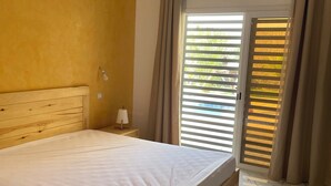 A bedroom with a wooden bed, a nightstand, a lamp, and a window with blinds. - Luxury villa with private pool and staff close to the beach and Saly (N'Guerigne Bambara)