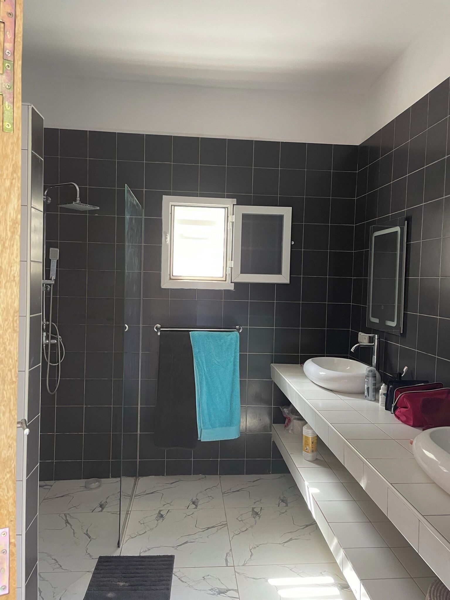 Un baño moderno con baldosas oscuras, cabina de ducha de vidrio, toallero con una toalla azul y tocador con dos lavamanos y artículos de aseo.