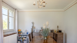 A dining room with a wooden table, chairs, a chandelier, a radiator, and a bookshelf. - Waterfront Apartment with Garden (Limoux)