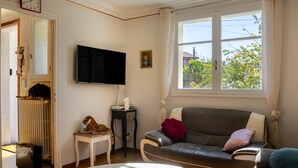 A living room with a sofa, a TV, a table, and a window with a view of trees. - Waterfront Apartment with Garden (Limoux)