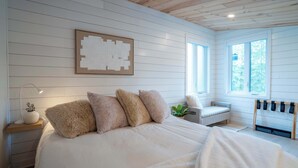 Een slaapkamer met een groot bed, een nachtkastje met een lamp, een stoel en een raam met uitzicht op bomen.