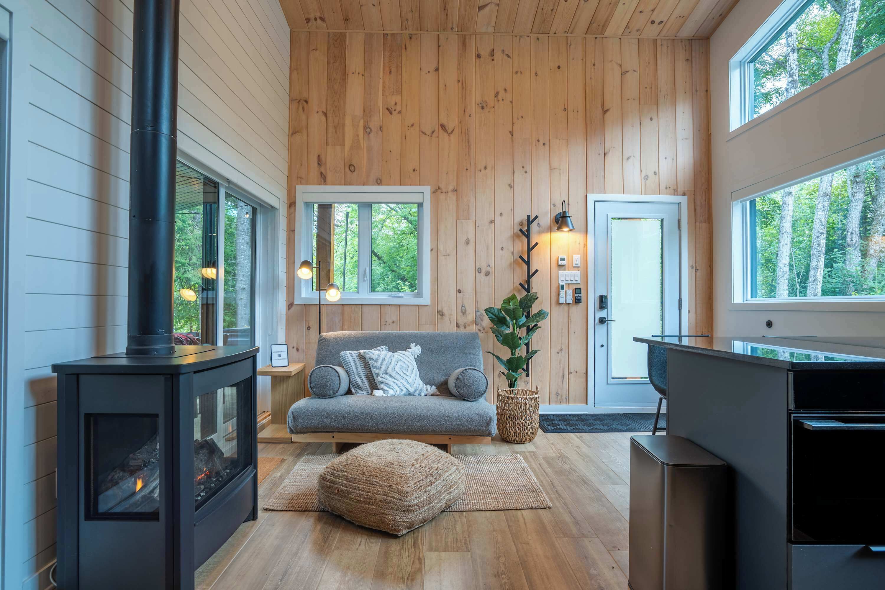 A cozy cabin interior with a wood-paneled ceiling, a sofa, a fireplace, and large windows offering views of the forest.