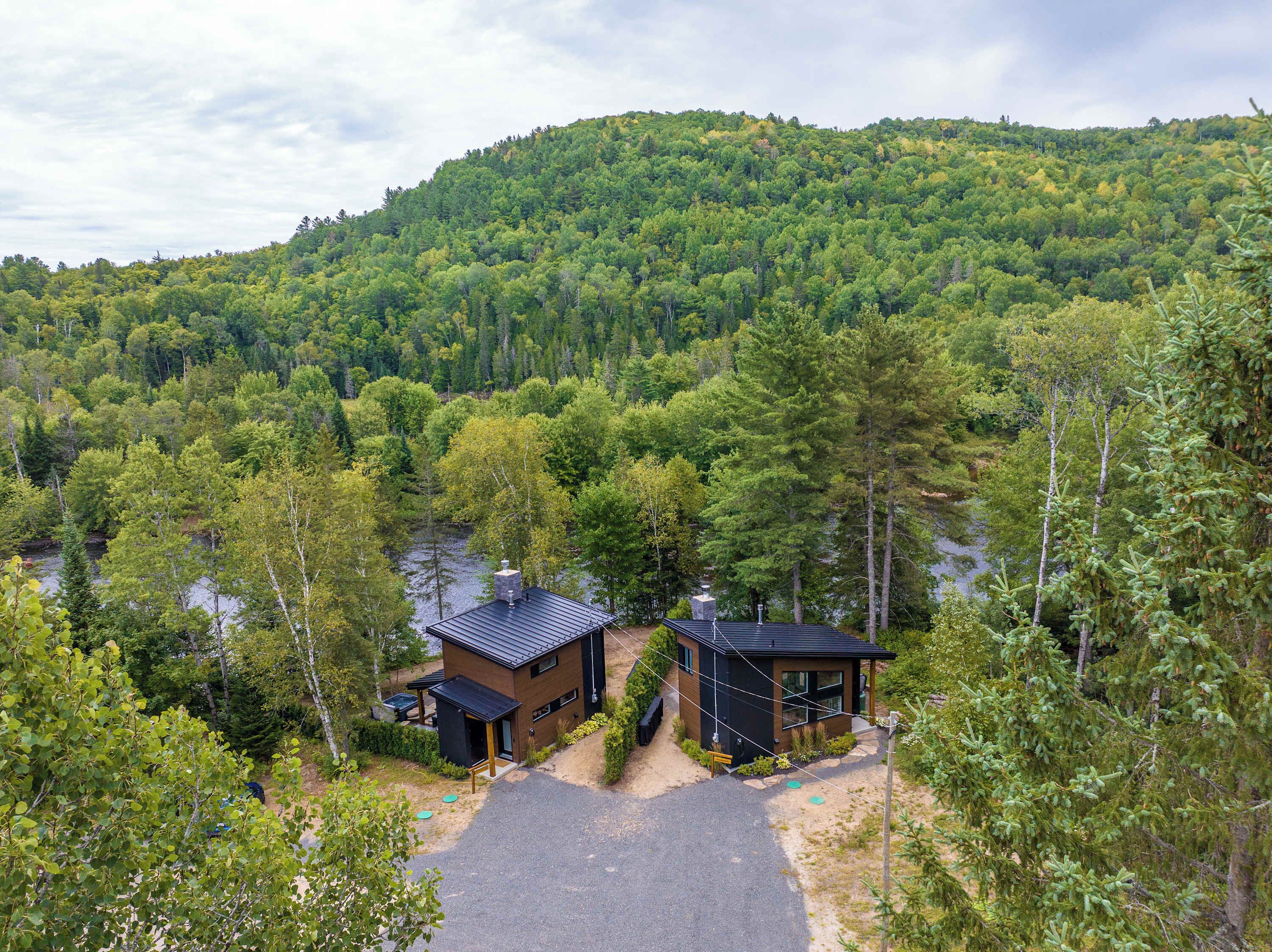 A cabin with a paved driveway and a small outdoor seating area, surrounded by dense forest and a lake.