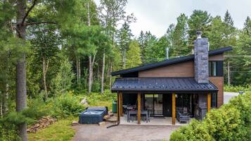 A modern house with a stone chimney, a covered patio, and a hot tub in a forested area.