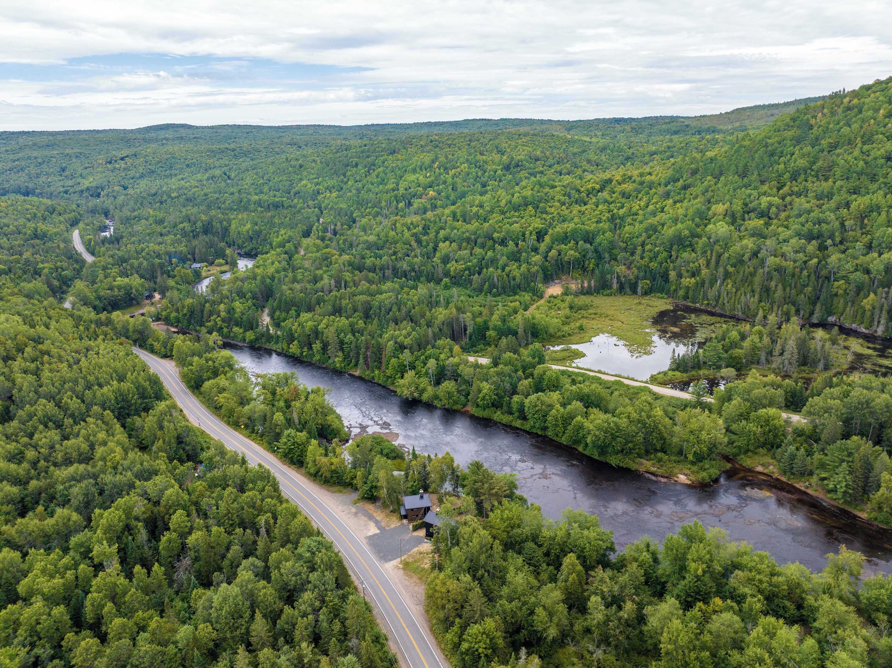 An aerial view of a forested area with a winding road, a small building near the road, and a river.
