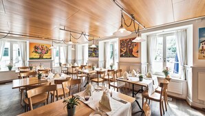 Un restaurant avec des tables et des chaises en bois, des tableaux accrochés aux murs et de grandes fenêtres.