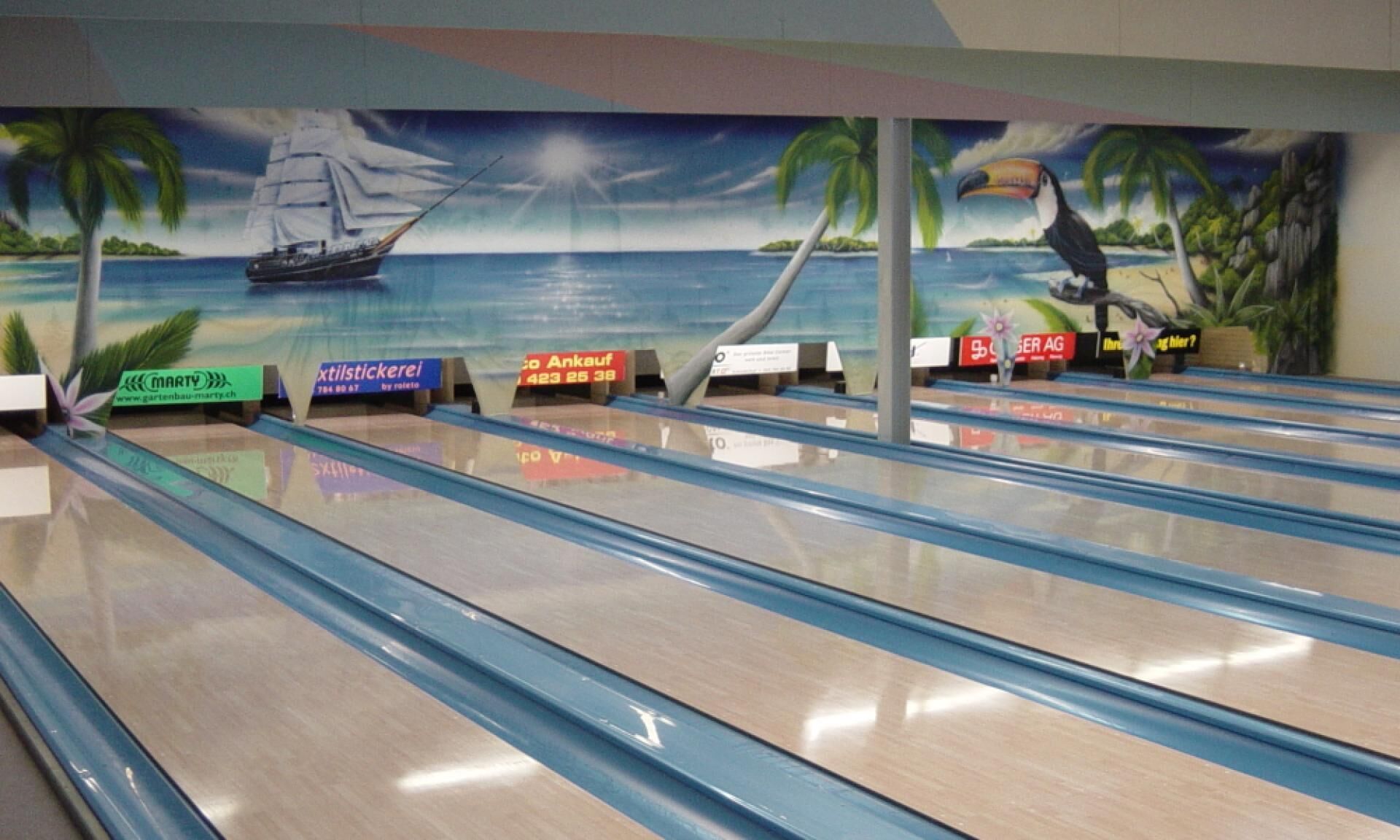 A bowling alley with lanes, a mural of a sailboat on water, palm trees, and a toucan.