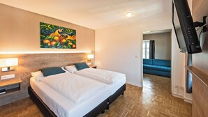 Une chambre d’hôtel avec un lit, deux tables de chevet, un tableau, une télévision et un canapé bleu dans la pièce adjacente.