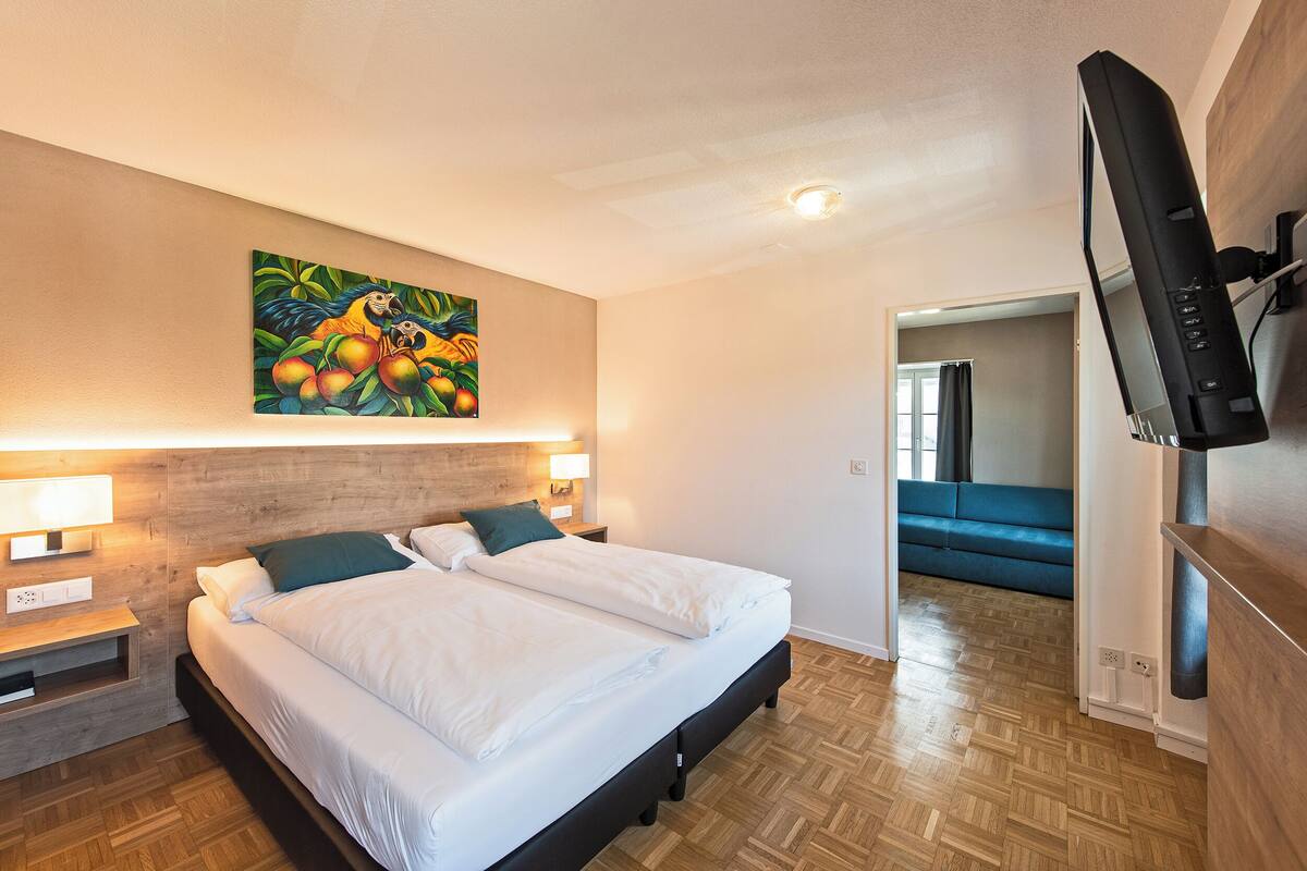 Une chambre d’hôtel avec un lit, deux tables de chevet, un tableau, une télévision et un canapé bleu dans la pièce adjacente.