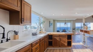 A modern kitchen with wooden cabinets, a white countertop, and a large window offering a view of the sea. - @ Marbella Lane - Ocean Views & Heavenly Sunset (Pacifica)