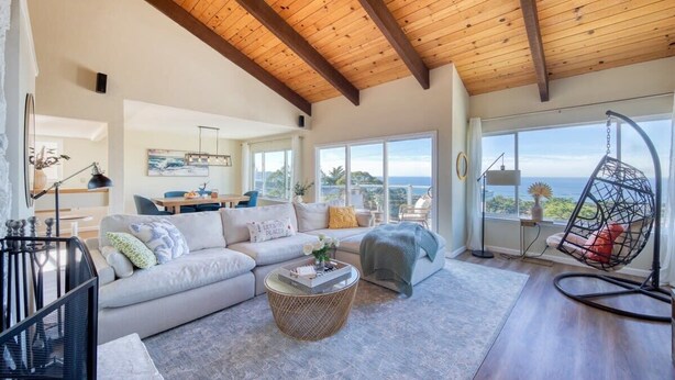 A spacious living room with a high ceiling, a large window offering a view of the outdoors, a cozy seating area with a sofa and a hanging chair, and a dining area in the background. - @ Marbella Lane - Ocean Views & Heavenly Sunset (Pacifica)