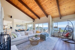 A spacious living room with a high ceiling, a large window offering a view of the outdoors, a cozy seating area with a sofa and a hanging chair, and a dining area in the background. - @ Marbella Lane - Ocean Views & Heavenly Sunset (Pacifica)