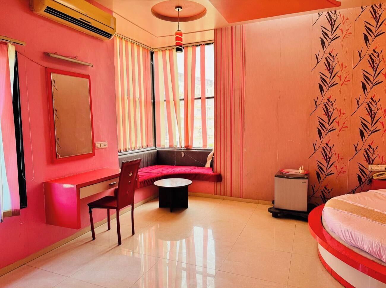 A room with a red and white color scheme, a bed, a desk with a chair, a small table, and a window with curtains.