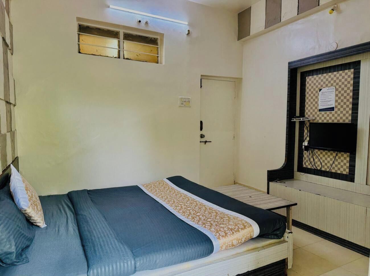 Un lit simple avec un matelas bleu et une couverture à motifs, une petite table de chevet, une fenêtre avec un store à carreaux et une porte avec une poignée.