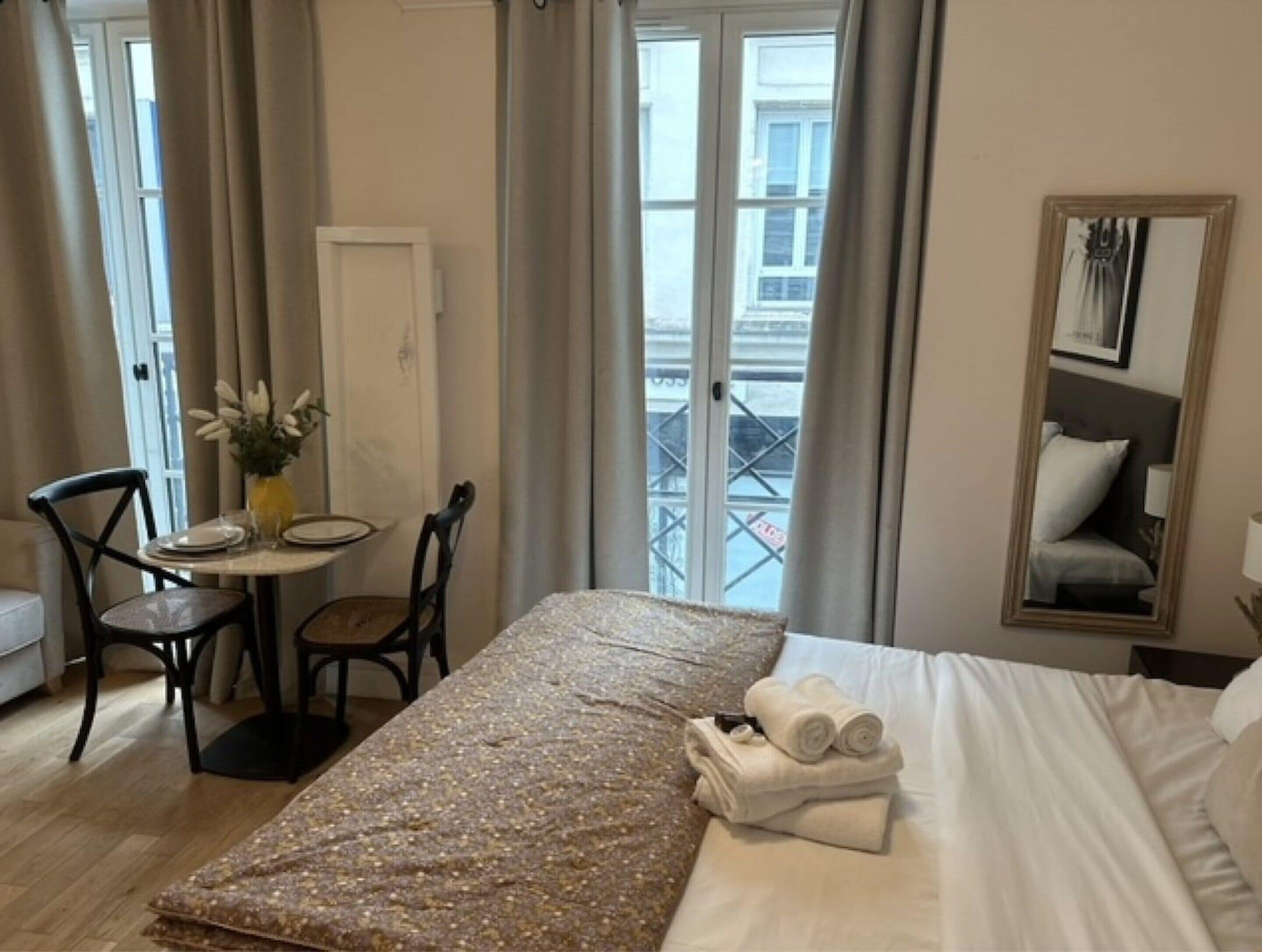 A hotel room with a bed, a small table with chairs, a vase with flowers, and a view of the outside through a window.