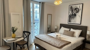 A hotel room with a bed, a desk with a chair, a small table with a vase, a mirror, and a framed picture on the wall. - Lovely Studio With AC - Center of Paris (Paris)