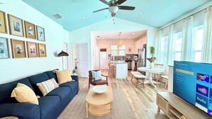 A spacious living room with a blue sofa, a wooden coffee table, and a TV on a stand. There is a ceiling fan, a kitchen in the background, and a dining area with a table and chairs. - The Royal Cypress Suite, Near UF, Shands, Downtown (Gainesville)