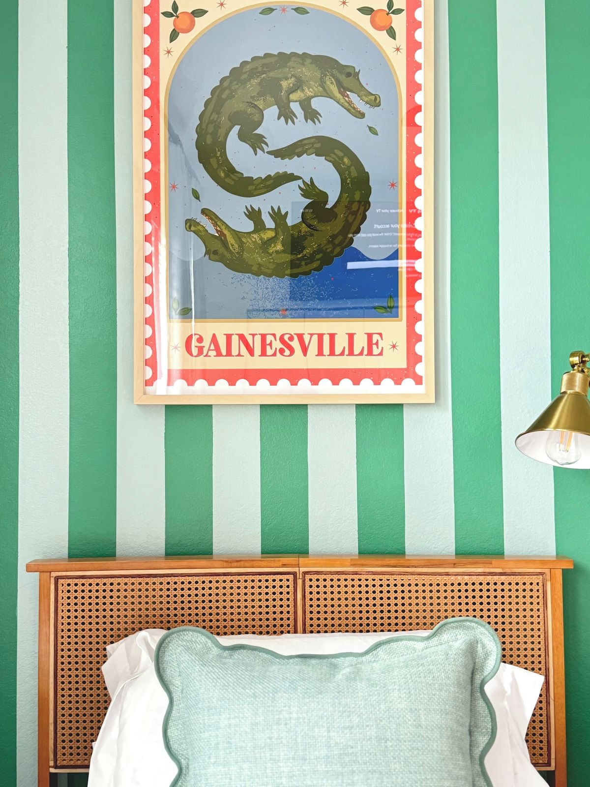 Une pièce avec un mur rayé de vert et de blanc, une affiche encadrée représentant un crocodile, avec le mot « GAINESVILLE » en dessous, une tête de lit en bois et une lampe sur la droite.