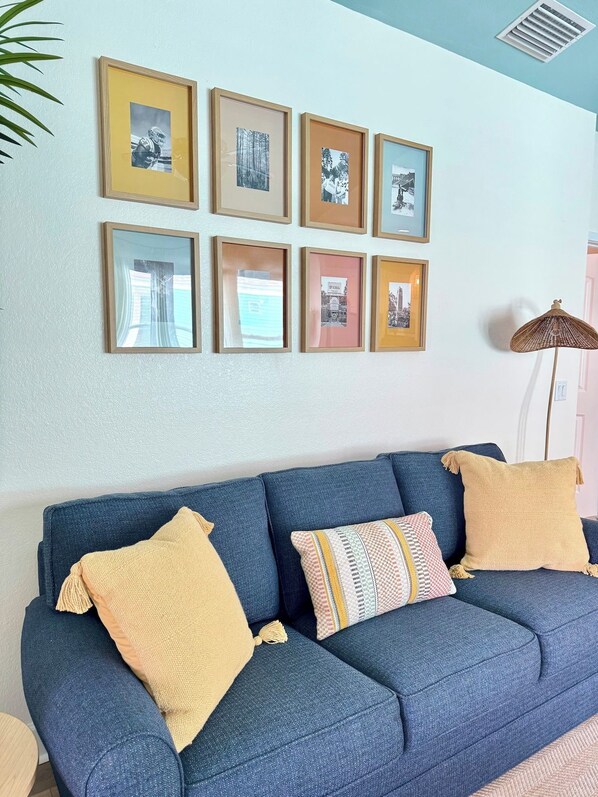 Un sofá azul con cojines amarillos y estampados, una pared con cuadros enmarcados, y una lámpara de pie.