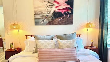 Une chambre d’hôtel avec un grand lit, deux tables de chevet, une petite table d’appoint, un tableau représentant un flamant rose et deux lampes fixées au mur.